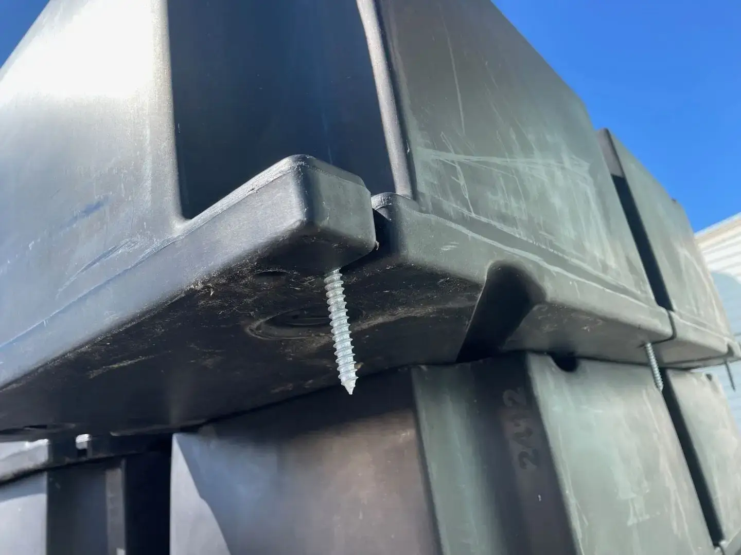 A screw is lodged in the bottom corner of a stacked black Dock Float Attachment Kit (Foam Filled) from Great Northern Docks, with clear blue sky behind.