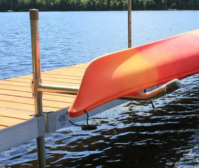 A red and orange kayak sits on a Kayak & Canoe Arm (Single Arm) by Great Northern Docks, mounted to a wooden dock by a calm lake with trees in the background.