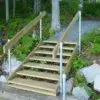 Metal rail brackets support wooden outdoor stairs that descend from a gravel path through greenery to a rocky area near Great Northern Docks.
