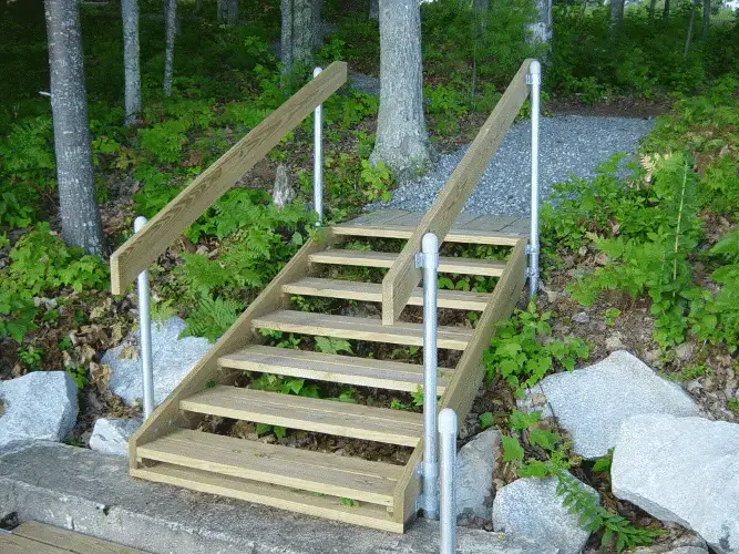 Metal rail brackets support wooden outdoor stairs that descend from a gravel path through greenery to a rocky area near Great Northern Docks.