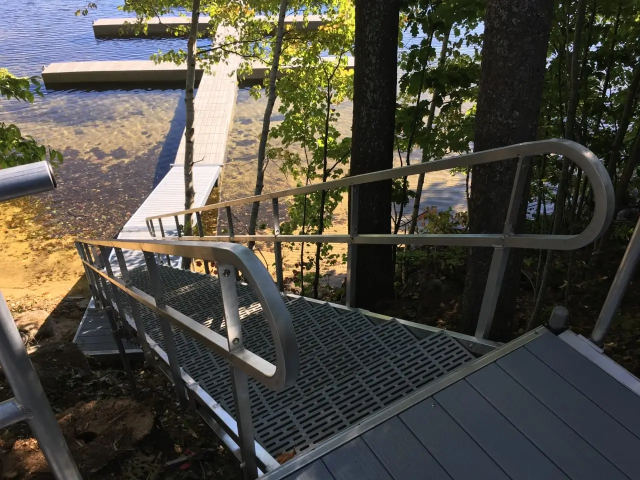 Fully Welded Stairs with railings lead through trees to a Maine dock over clear, shallow water, with sunlight streaming through the leaves.