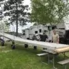 Three men stand outdoors with a Dock Wheel Kit from Great Northern Docks near a picnic table, RV, and trees by a lake.