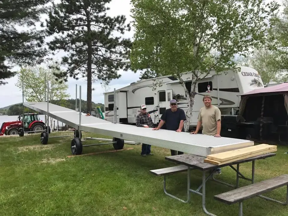Three men stand outdoors with a Dock Wheel Kit from Great Northern Docks near a picnic table, RV, and trees by a lake.