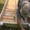 A person wearing a straw hat assembles a wooden frame for Wood Docks outdoors, using tools and metal brackets on a grassy surface.
