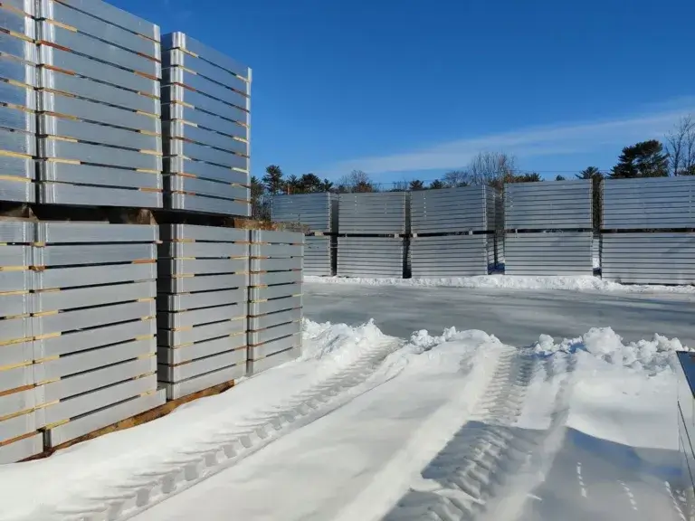 Stacks of metal sheets are arranged outdoors on snowy ground near Great Northern Docks, with visible tire tracks and a clear blue sky in the background.