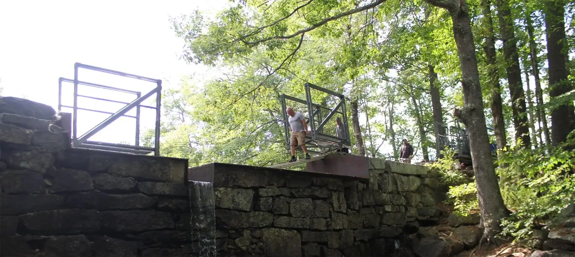 A person stands on an Arched Footbridge by an open metal gate, with water flowing over the edge, surrounded by trees and Maine Docks.