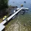 Aerial view of Boarding Steps on Maine docks extending into a clear lake, with boats, jet skis, and a floating play structure nearby; tree-lined rocky shore.