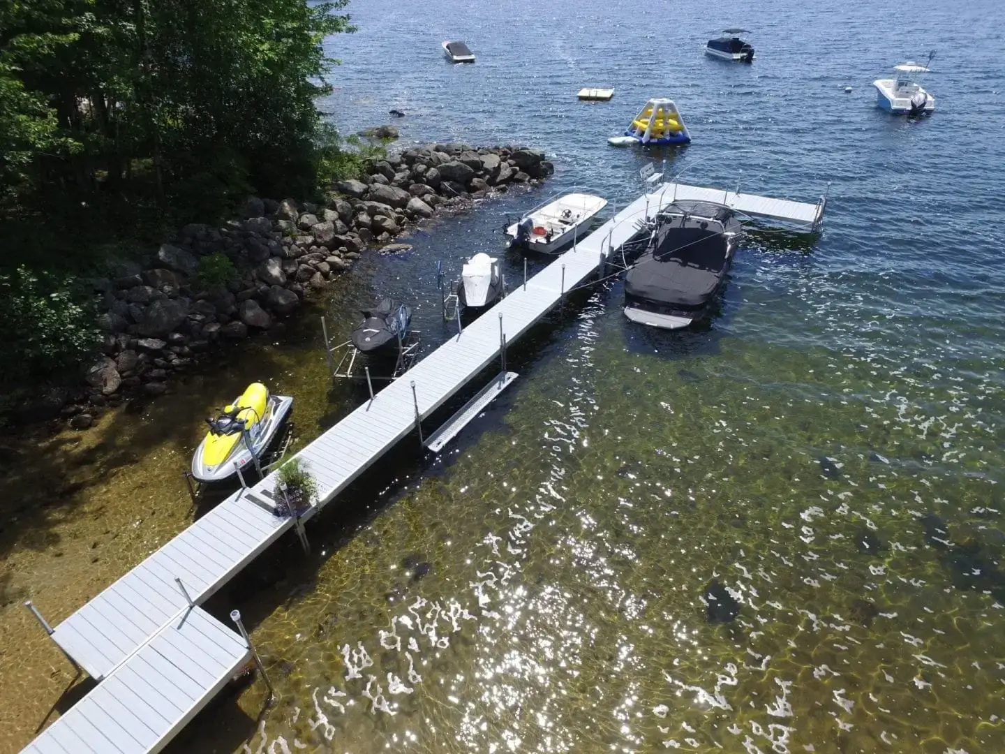 Aerial view of Boarding Steps on Maine docks extending into a clear lake, with boats, jet skis, and a floating play structure nearby; tree-lined rocky shore.