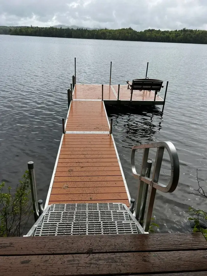 A wooden dock with metal railings extends over a calm lake, ending at a platform with a bench. Wood Docks blend seamlessly with the trees and cloudy sky in the background.