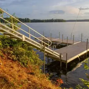 A wooden dock by Great Northern Docks with sturdy railings extends from a grassy shore into a calm lake, framed by trees and a partly cloudy sunset sky.