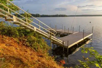 A wooden dock by Great Northern Docks with sturdy railings extends from a grassy shore into a calm lake, framed by trees and a partly cloudy sunset sky.
