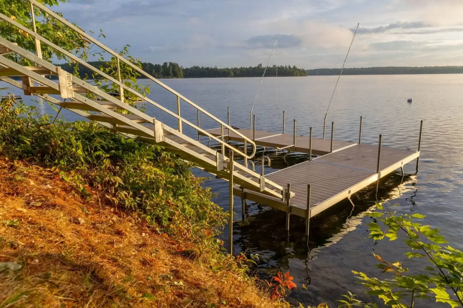 A wooden dock by Great Northern Docks with sturdy railings extends from a grassy shore into a calm lake, framed by trees and a partly cloudy sunset sky.