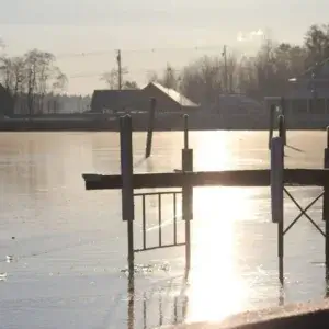 A wooden dock from Great Northern Docks extends over a frozen lake, reflecting sunlight. Houses and trees are visible in the background on a cold, clear day.