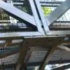 Close-up of the Arched Footbridge's steel structure with intersecting beams and metal grating near Great Northern Docks, trees and sunlight in the background.