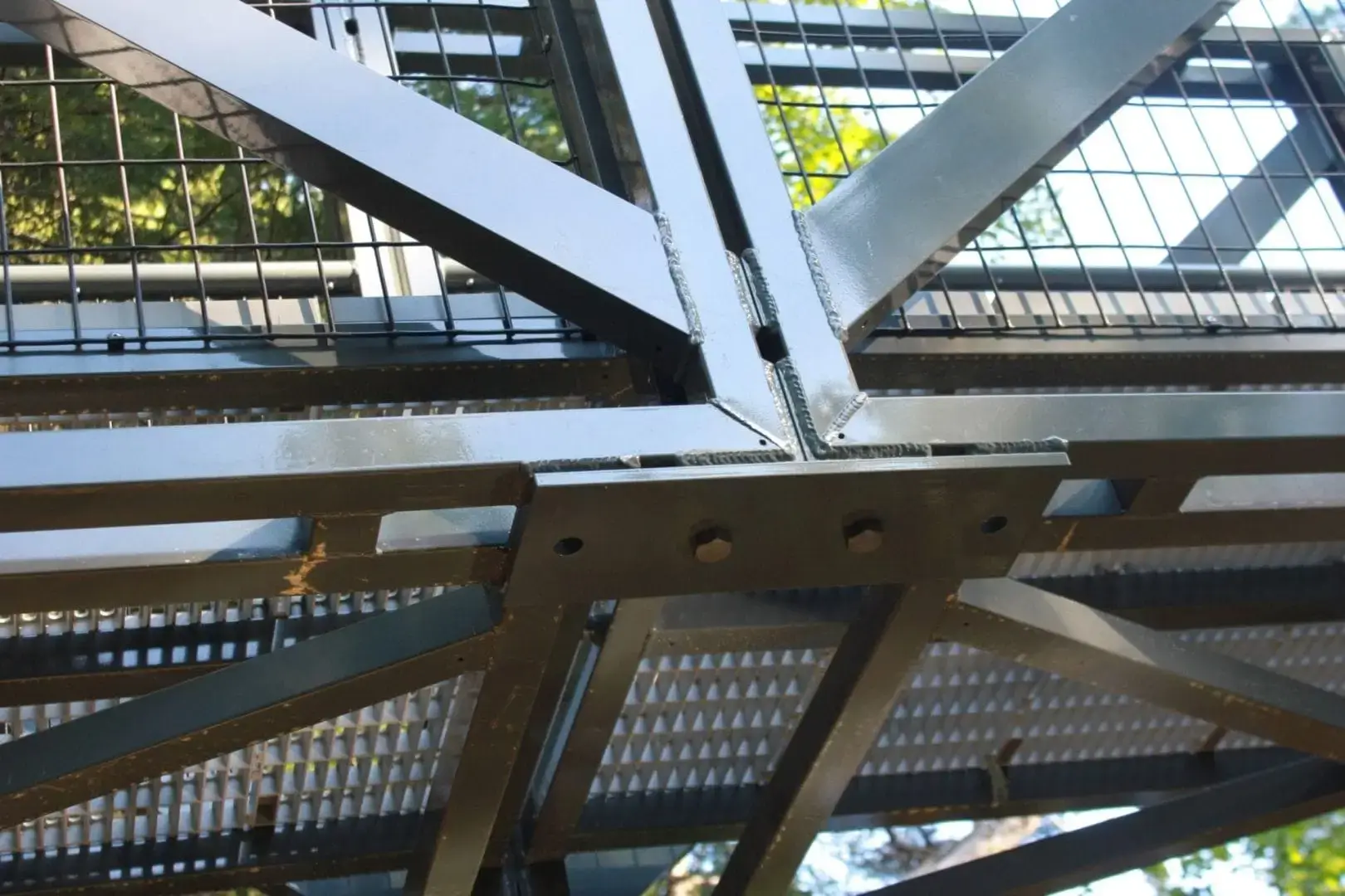 Close-up of the Arched Footbridge's steel structure with intersecting beams and metal grating near Great Northern Docks, trees and sunlight in the background.