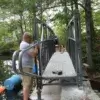 A person assembles an Arched Footbridge over a creek in the woods, using tools on rocks—like constructing sturdy Wood Docks from Great Northern Docks.