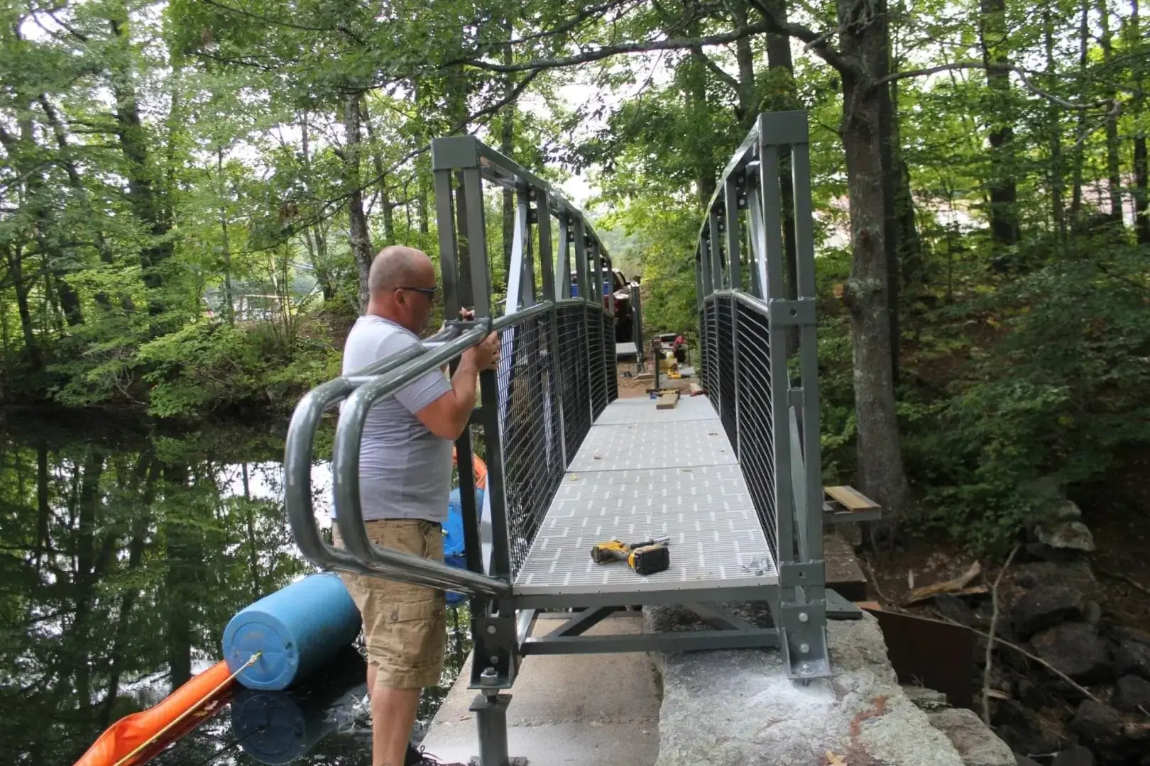 A person assembles an Arched Footbridge over a creek in the woods, using tools on rocks—like constructing sturdy Wood Docks from Great Northern Docks.