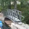 An Arched Footbridge spans a narrow canal in the woods, evoking classic Maine Docks scenes as someone works nearby.