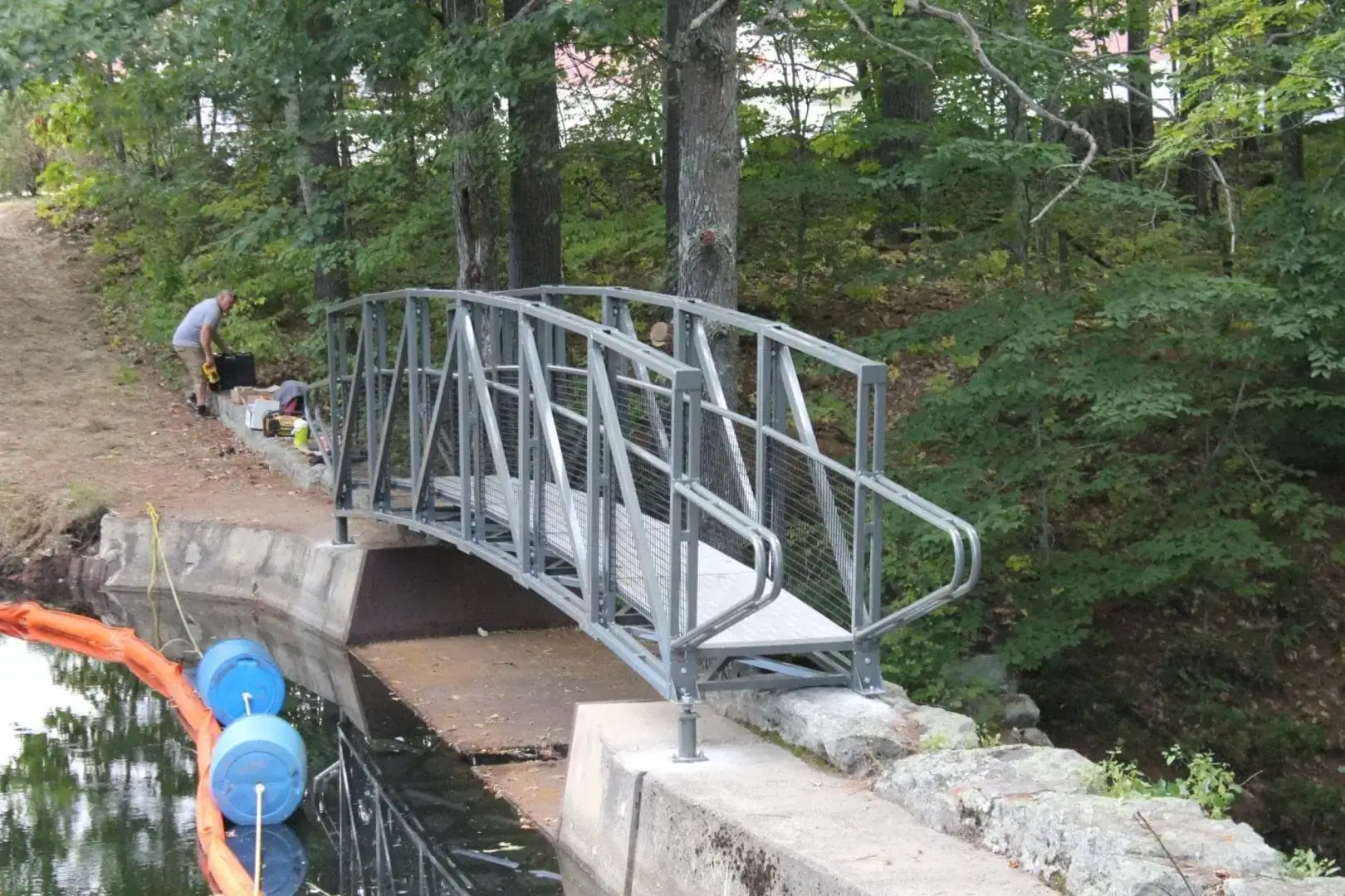 An Arched Footbridge spans a narrow canal in the woods, evoking classic Maine Docks scenes as someone works nearby.