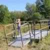 Two people stand on a metal Boardwalks structure near Maine Docks in a grassy field, using binoculars. Trees and blue sky complete the scene.