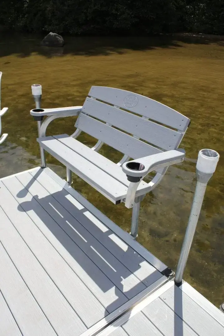 A Bench with armrests and cup holders is installed on a Great Northern Docks wood dock above shallow water near trees.