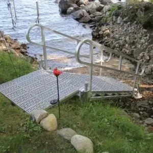 A metal ramp with railings leads down a grassy slope to rocky lakeshore docks, flanked by two torches and water shimmering in the background.