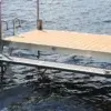 A floating Maine Dock with a tan deck and metal frame extends over rippling water, featuring Boarding Steps attached at the end.