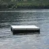 A small Swim Raft with a metal ladder floats on calm lake water, surrounded by trees along the distant shore.