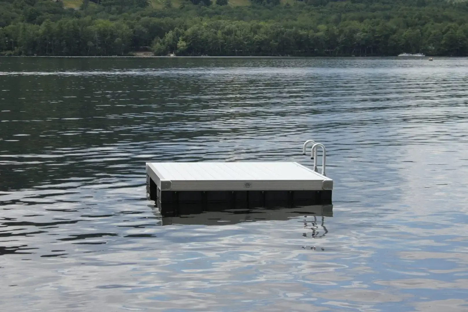 A small Swim Raft with a metal ladder floats on calm lake water, surrounded by trees along the distant shore.