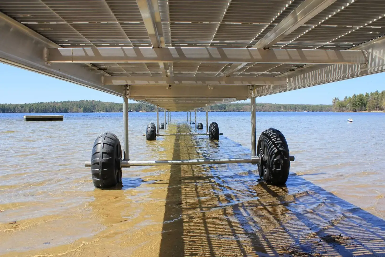 View from under a Roller Dock with large black wheels over clear shallow water, extending toward a lake, with Maine Docks and trees in the background.