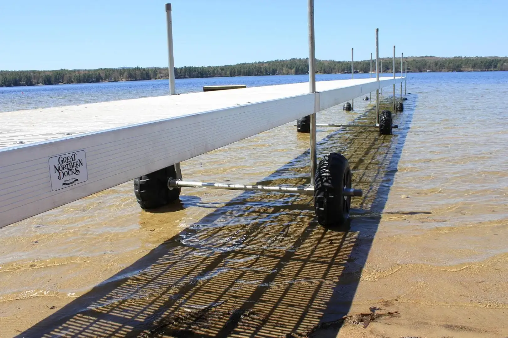 The Roller Dock from Great Northern Docks extends on wheels into a clear, shallow lake with a sandy bottom and trees lining the distant shore under a blue sky.