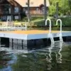 A Swim Raft with a metal ladder floats on a calm lake, with Maine docks and tree-lined houses visible on the distant shoreline.