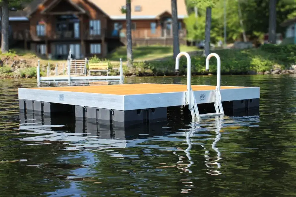 A Swim Raft with a metal ladder floats on a calm lake, with Maine docks and tree-lined houses visible on the distant shoreline.