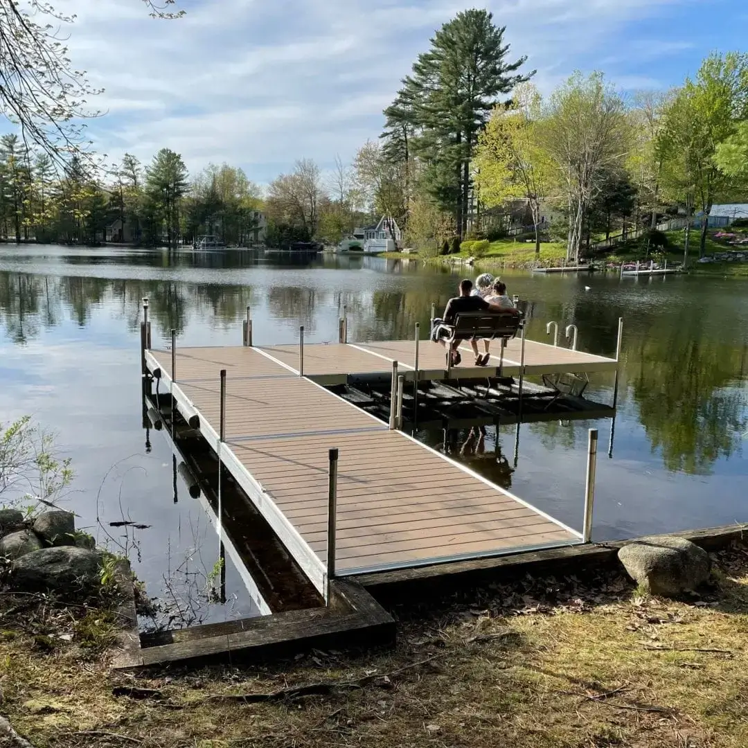 A Bench is placed at the end of a wooden dock over a calm lake, surrounded by trees and cabins under sunny skies.