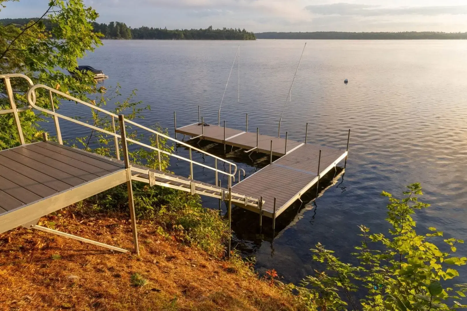 A metal ramp leads down to an Aluminum Dock on a calm lake, surrounded by trees, with sunlight reflecting on the water in the early morning or evening.
