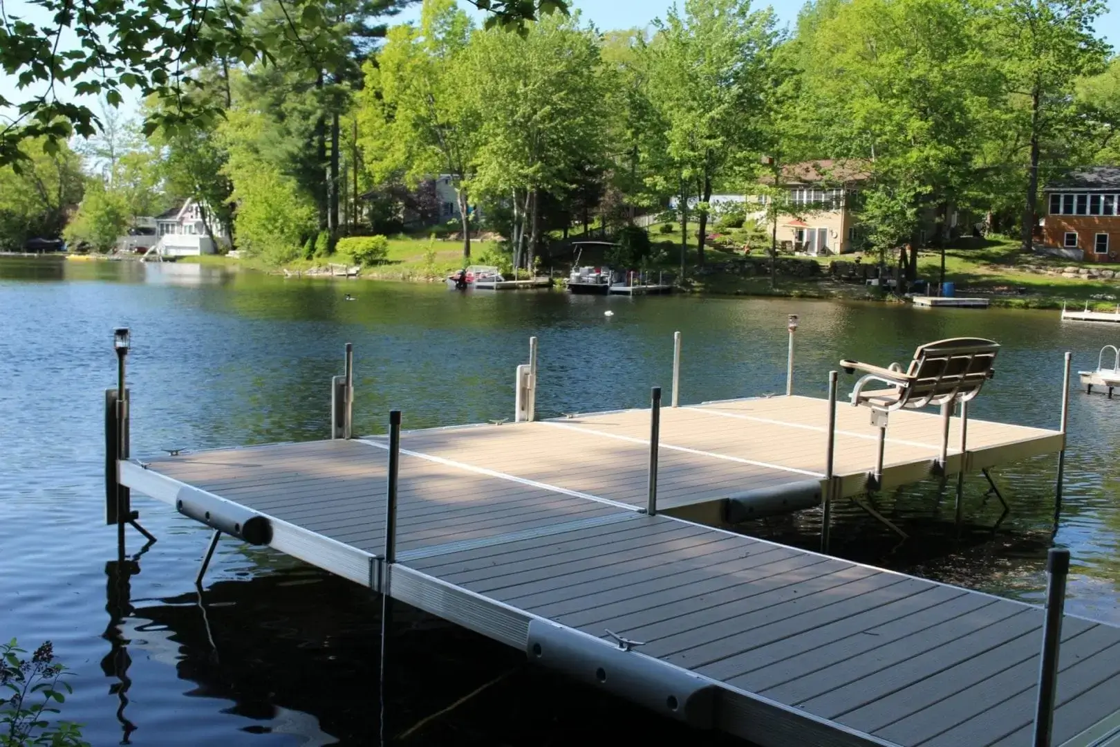 A wooden dock by Great Northern Docks with benches extends over a calm lake, surrounded by trees and lakeside houses under a clear blue sky.