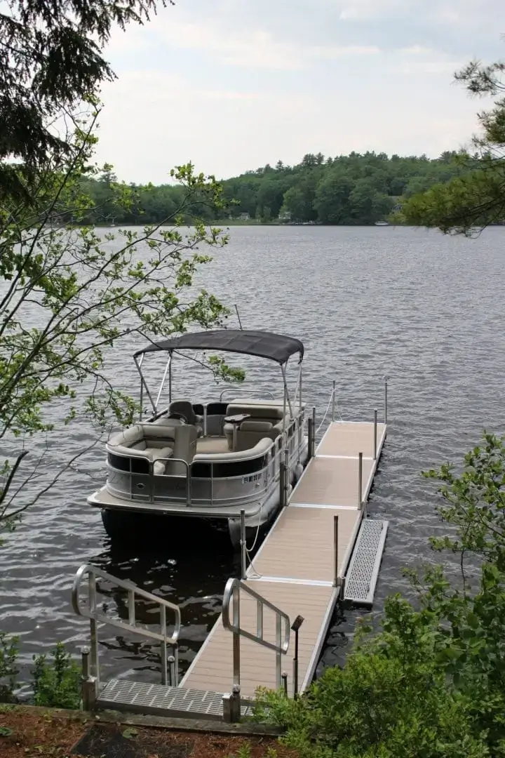 A pontoon boat is docked at a small pier with classic wood docks on a calm lake, surrounded by trees and greenery under a partly cloudy sky.