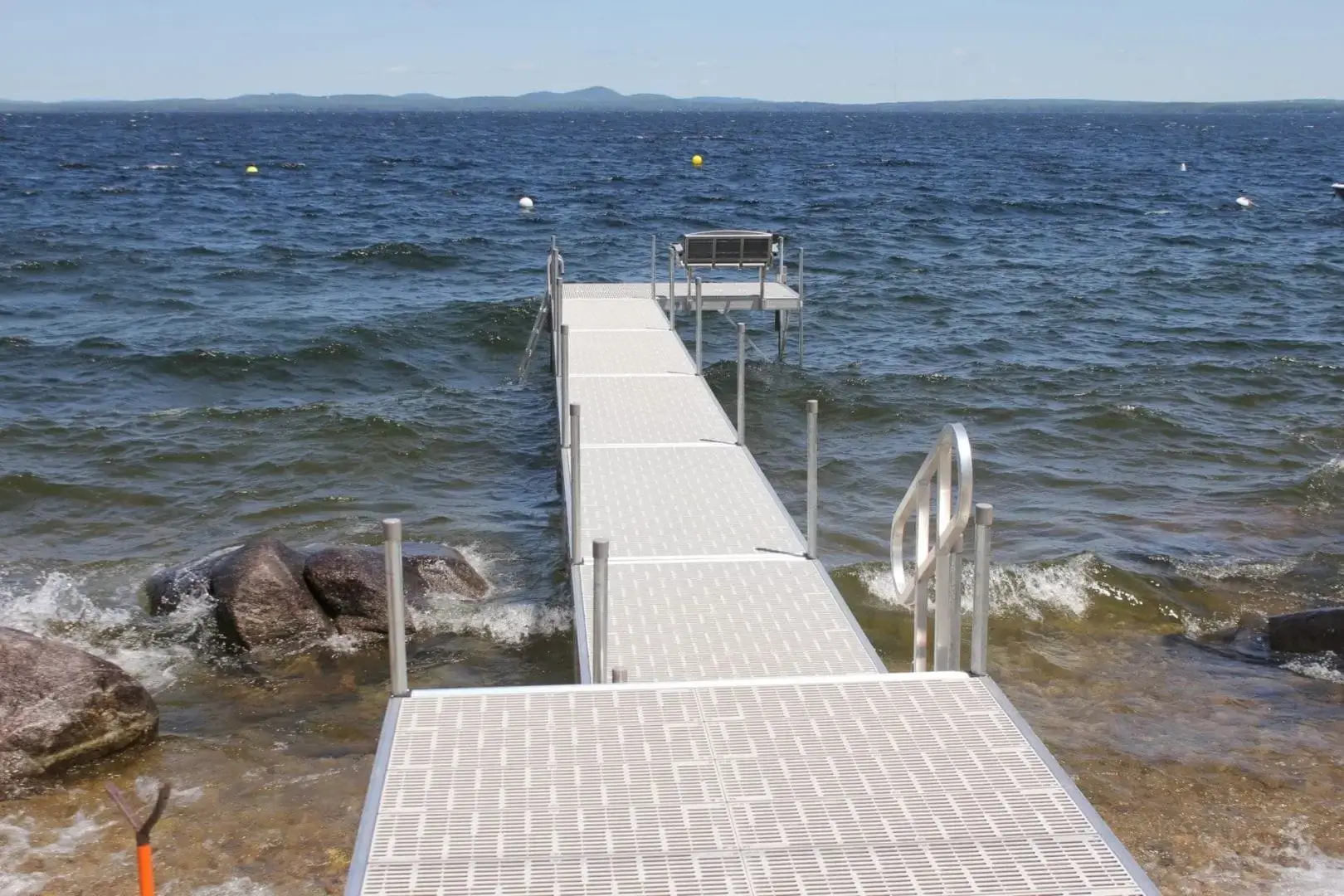 A Great Northern Docks aluminum dock extends into a choppy lake, with rocks on the shore and a bench at the end under a clear blue sky.