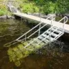 A metal ramp with handrails extends from a wooden dock by Maine Docks into the water, allowing easy access for walking or wheelchairs. Green foliage and rocks line the shore.