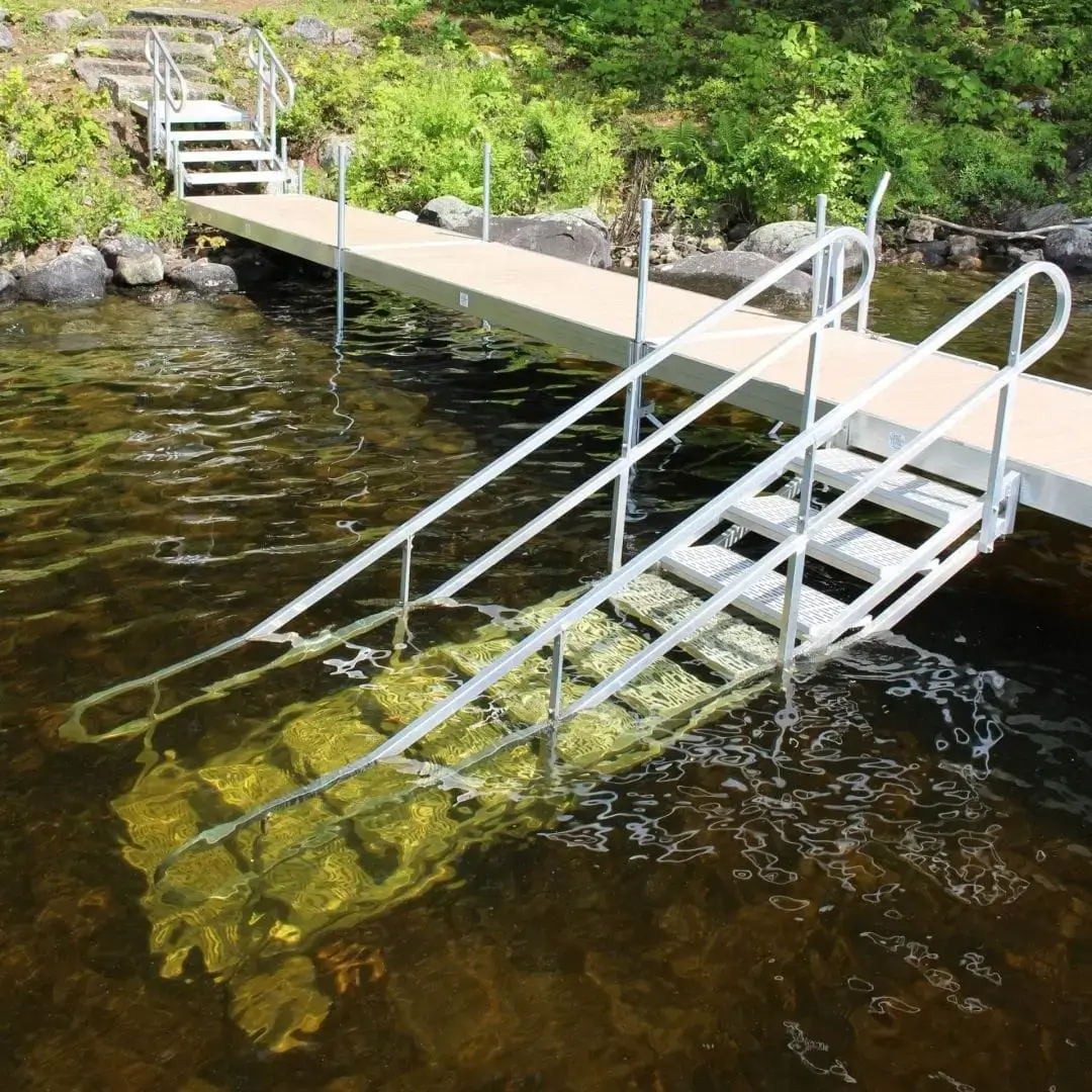 A metal ramp with handrails extends from a wooden dock by Maine Docks into the water, allowing easy access for walking or wheelchairs. Green foliage and rocks line the shore.