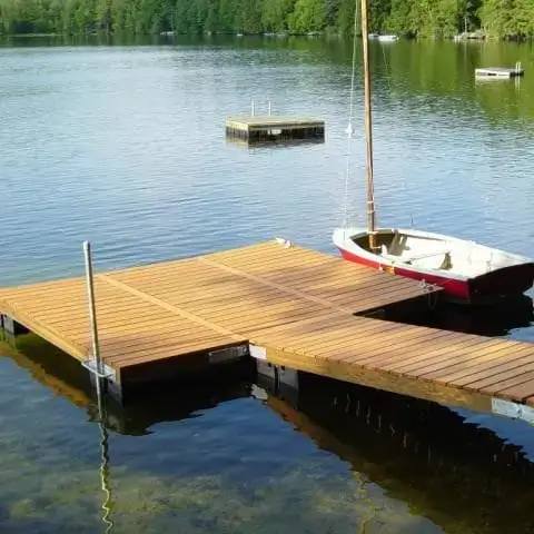 A wooden dock extends into a calm lake with a small red and white sailboat tied to it. Trees line the far shore, and a floating platform highlights classic Maine Docks scenery.