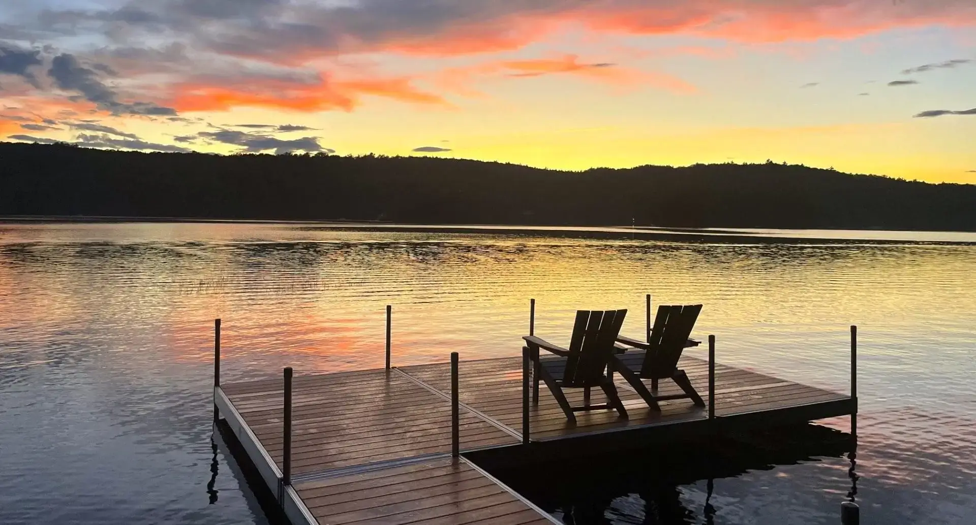 Two empty chairs sit on a Wood Dock by Great Northern Docks, overlooking a calm lake at sunset, with colorful clouds and silhouetted trees in the background.