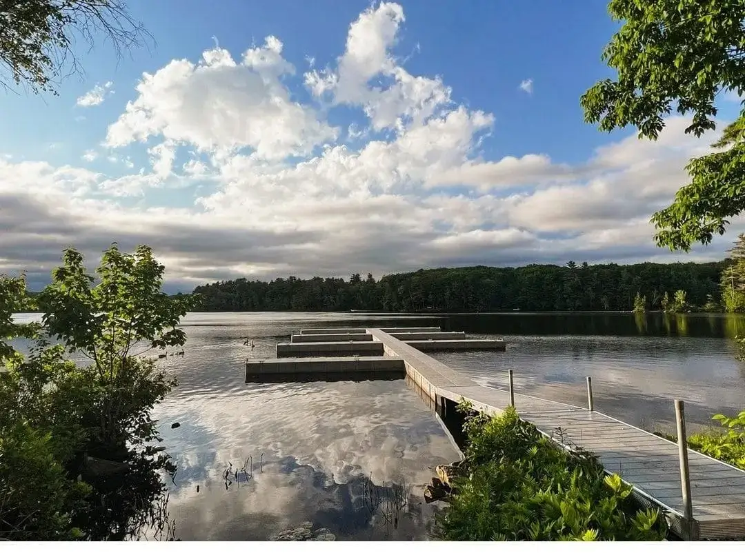 A wooden dock from Great Northern Docks extends over a calm lake, surrounded by green trees beneath a partly cloudy blue sky.
