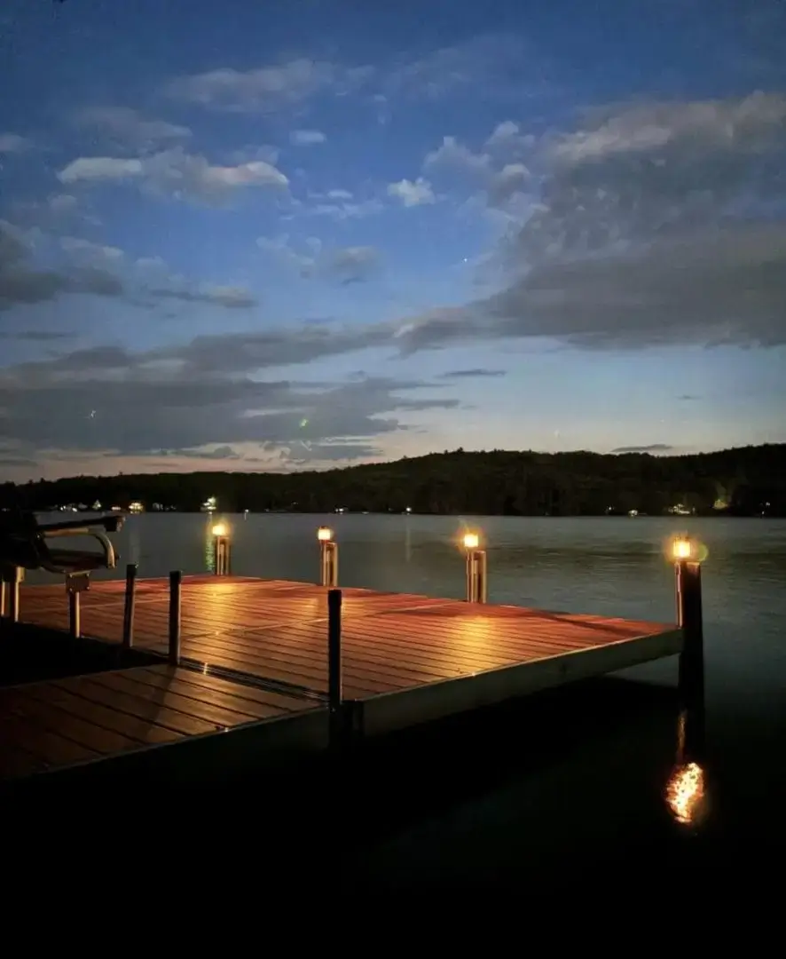 A wooden dock with lights glows at dusk, overlooking a calm lake under a partly cloudy sky with trees in the distance.