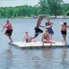 1000Swim-low Six people enjoy a sunny day on a Swim Raft - Connect-A-Dock; some leap into the lake while others relax and smile, surrounded by Maine's scenic trees.