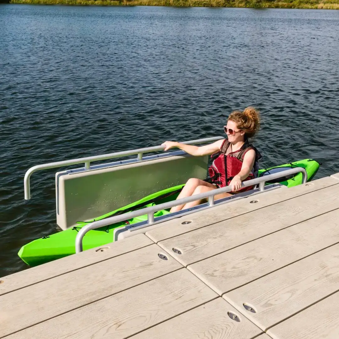 3 A woman in a green kayak uses a Connect-A-Dock Kayak Launch to enter the water, wearing sunglasses and a life jacket on a sunny day.