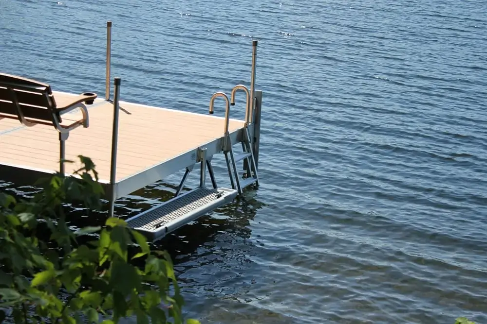 A metal and wood dock with a bench and ladder extends over calm blue lake water, with some green foliage visible in the foreground.