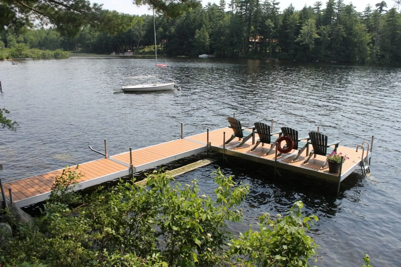A wooden dock with four Adirondack chairs and a flower pot extends over a calm lake, with a sailboat anchored near the docks and trees lining the shore.