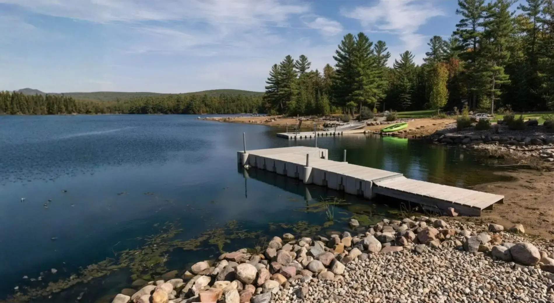 A wooden dock extends into a calm lake bordered by rocks and trees, with another Great Northern Docks kayak dock and a green kayak visible on the shore under a blue sky.