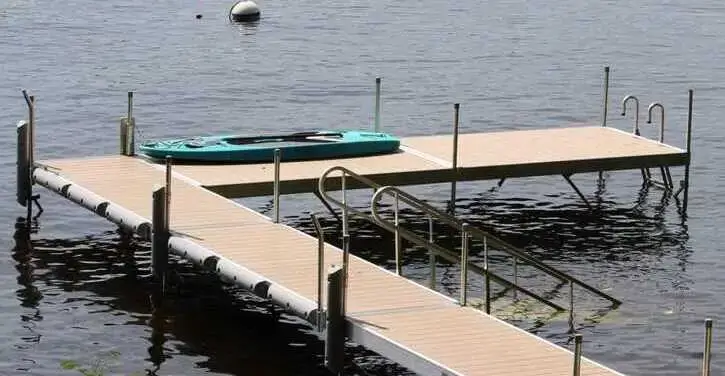 A wooden dock extends over a calm lake, with a teal kayak resting on it and a white buoy floating in the water nearby.
